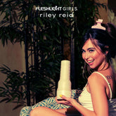 A woman with a hair clip smiles at the camera while holding the Fleshlight Girls: Riley Reid Utopia by Fleshlight, featuring lifelike SuperSkin. She sits indoors with a leafy plant in the background. The text reads "Fleshlight Girls Riley Reid Utopia.
