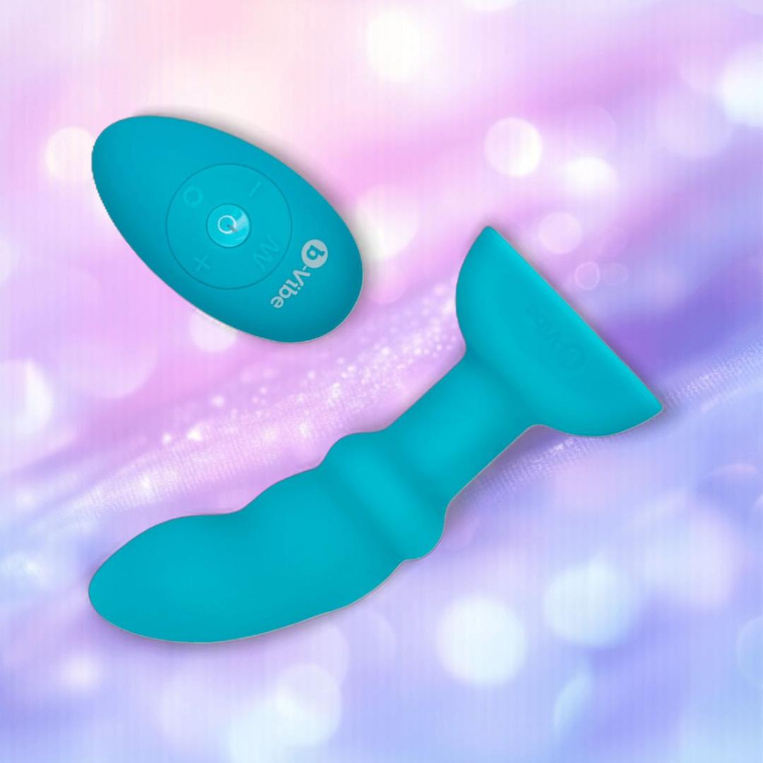 The B-Vibe Remote Rimming P-Spot Plug by B-Vibe, a blue silicone vibrating anal plug with remote control, is shown on a pastel purple and pink bokeh background. The B-Vibe Remote Rimming P-Spot Plug by B-Vibe, a blue silicone vibrating anal plug with remote control, is shown on a pastel purple and pink bokeh background.