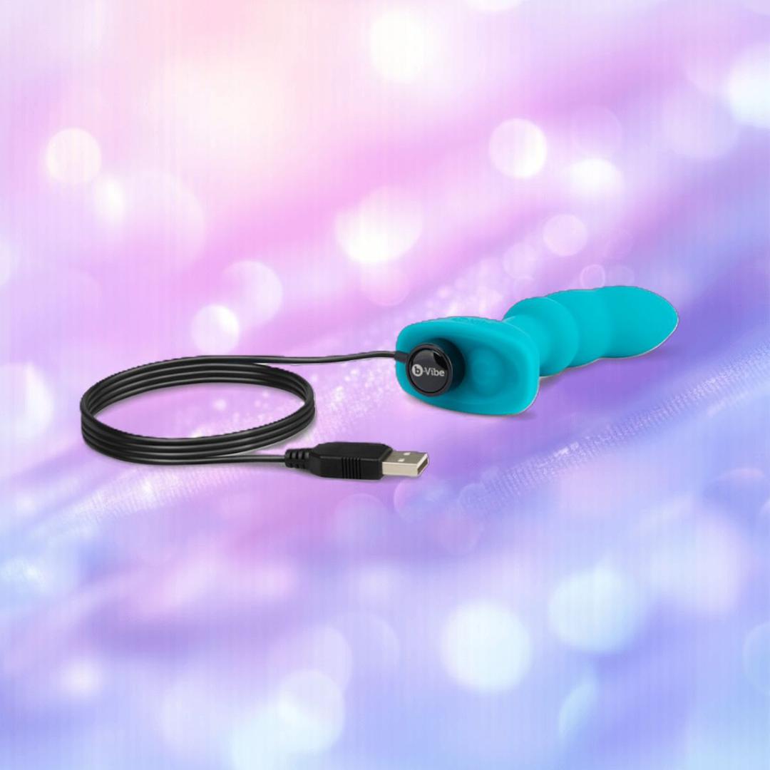 The B-Vibe Remote Rimming P-Spot Plug by B-Vibe, a teal silicone vibrator with USB charging cable, is displayed on a soft pink and purple bokeh background. The B-Vibe Remote Rimming P-Spot Plug by B-Vibe, a teal silicone vibrator with USB charging cable, is displayed on a soft pink and purple bokeh background.