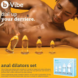 A yellow-themed image displays the B-Vibe Glass Anal Dilators Set of 4 in increasing sizes on a reflective surface. A person stands in the background with one hand on their hip, while text highlights b-Vibe's set for accessible and enjoyable anal training.