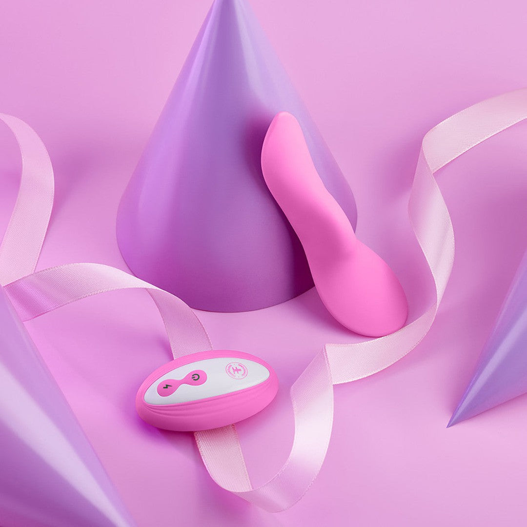 A pink and purple background showcases a small pink remote control and the matching FemmeFunn Unda Panty Vibrator in pink, featuring an ergonomic design. These are placed among pastel purple party hats and a white ribbon, creating a playful and festive atmosphere.