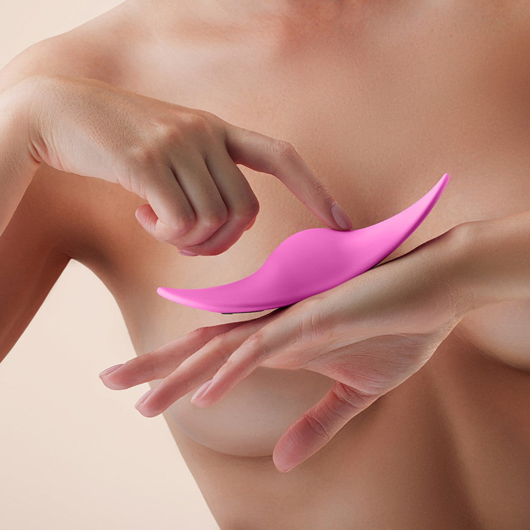 A person is holding a FemmeFunn Unda Panty Vibrator Pink at chest level. Their hands gently grip the ergonomic design, with fingers slightly curved around it. The background is a neutral beige.