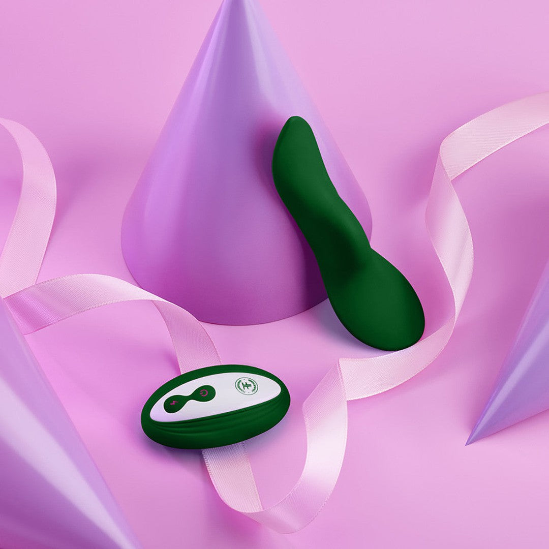 A FemmeFunn Unda Panty Vibrator in dark green and its remote control sit on a pink surface adorned with pastel purple party hats and a pink ribbon. The remote, featuring buttons and an LED indicator for discreet pleasure, adds to the playful scene with its soft, contrasting colors.