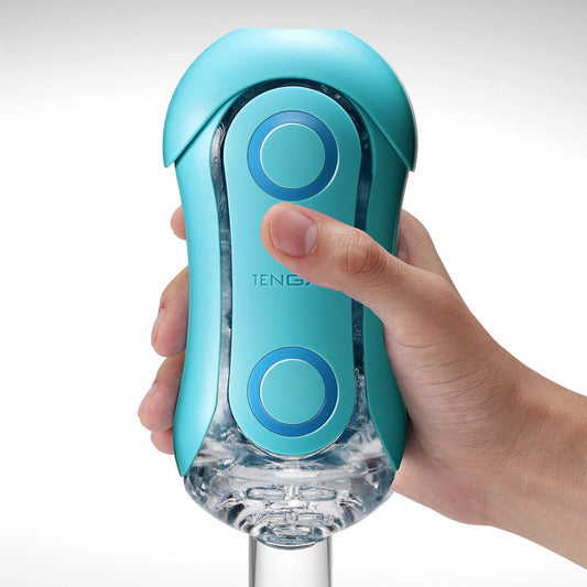 A hand holds a turquoise-colored Tenga Flip Orb Ocean Blue, a portable electronic masturbator featuring two blue circular buttons and a semi-transparent section revealing mechanical parts inside. The branding "TENGA" is clearly visible on the device. The background is neutral gray. 1080