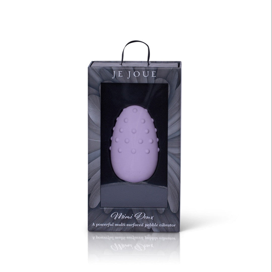 The Je Joue Mimi Deux Pebble Vibrator in Lilac is elegantly presented in a sleek black box with a transparent front. The brand name "Je Joue" is prominently displayed at the top, while the description "Mimi: A powerful multi-surfaced pebble vibrator" at the bottom emphasizes its dual-texture design.