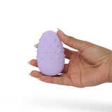 A hand gracefully holds the Je Joue Mimi Deux Pebble Vibrator in lilac, an oval-shaped object adorned with raised bumps on its surface, showcasing its unique dual-texture design against a plain white background.