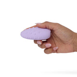 A hand holding a small, lilac, oval-shaped object with raised dots showcases the Je Joue Mimi Deux Pebble Vibrator's dual-texture design against a plain white background.