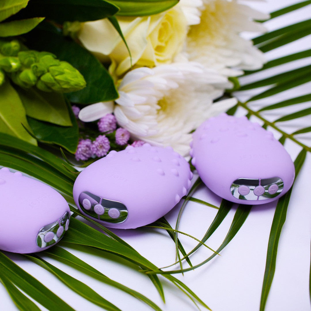 Three Je Joue Mimi Deux Pebble Vibrators in lilac, with a dual-texture design and low-frequency motor, featuring buttons on their ends, are placed on green palm leaves alongside white and purple flowers.