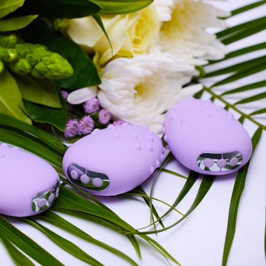 Three Je Joue Mimi Deux Pebble Vibrators in lilac, with a dual-texture design and low-frequency motor, featuring buttons on their ends, are placed on green palm leaves alongside white and purple flowers. 1080