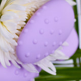 A close-up of the Je Joue Mimi Deux Pebble Vibrator in lilac, showcasing its elegant dual-texture design as it rests on white chrysanthemum petals. The backdrop features green leaves to enhance the fresh and botanical ambiance.