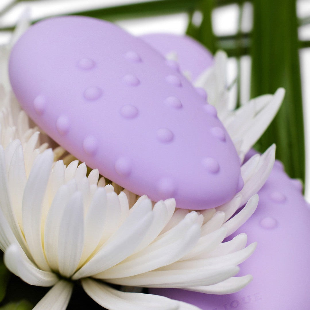 A Je Joue Mimi Deux Pebble Vibrator in lilac, featuring an oval shape with a dual-texture design and a dotted surface, rests charmingly on white flowers, set against a serene backdrop of green foliage.