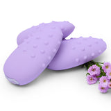 Three lilac Je Joue Mimi Deux Pebble Vibrators are stacked together on a white background, highlighting their dual-texture design. Small clusters of light pink flowers are placed beside them.