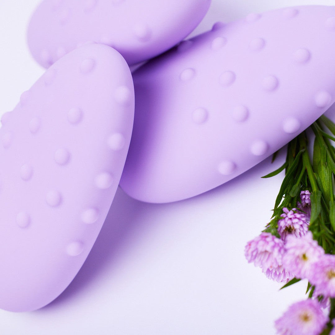 Close-up of three Je Joue Mimi Deux Pebble Vibrators in lilac, showcasing their dual-texture design and dot pattern, arranged beside a sprig of small purple flowers. The pastel purple tones against the white background evoke a soft, delicate composition characteristic of Je Joue elegance.