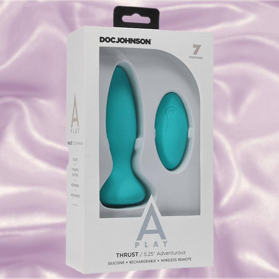 The A-Play Thrust Adventurous Rechargeable Plug in teal by Doc Johnson comes in white "A-Play" packaging and is shown with a wireless remote control against a light purple satin background.
