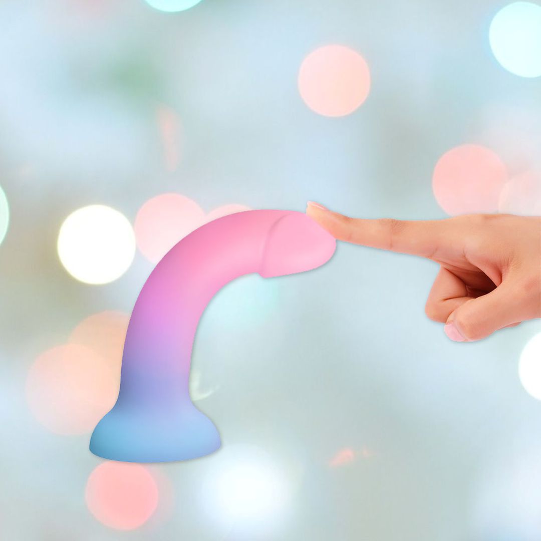 A human hand lightly touches the tip of a colorful DilDolls "Utopia" Silicone Dildo with Suction Base, featuring a gradient from pink to blue, against a softly blurred background with pastel bokeh lights.