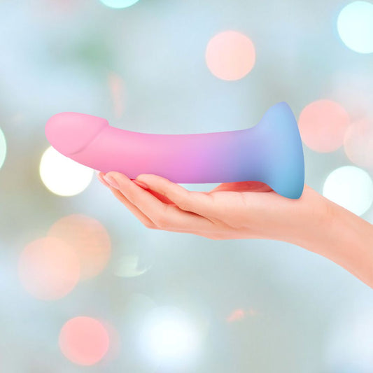 In a softly blurred, pastel bokeh background, a hand holds the Dildolls "Utopia" Silicone Dildo with Suction Base, featuring a gradient color that transitions from blue at the base to pink at the tip. This waterproof dildo combines both beauty and function seamlessly. 1080