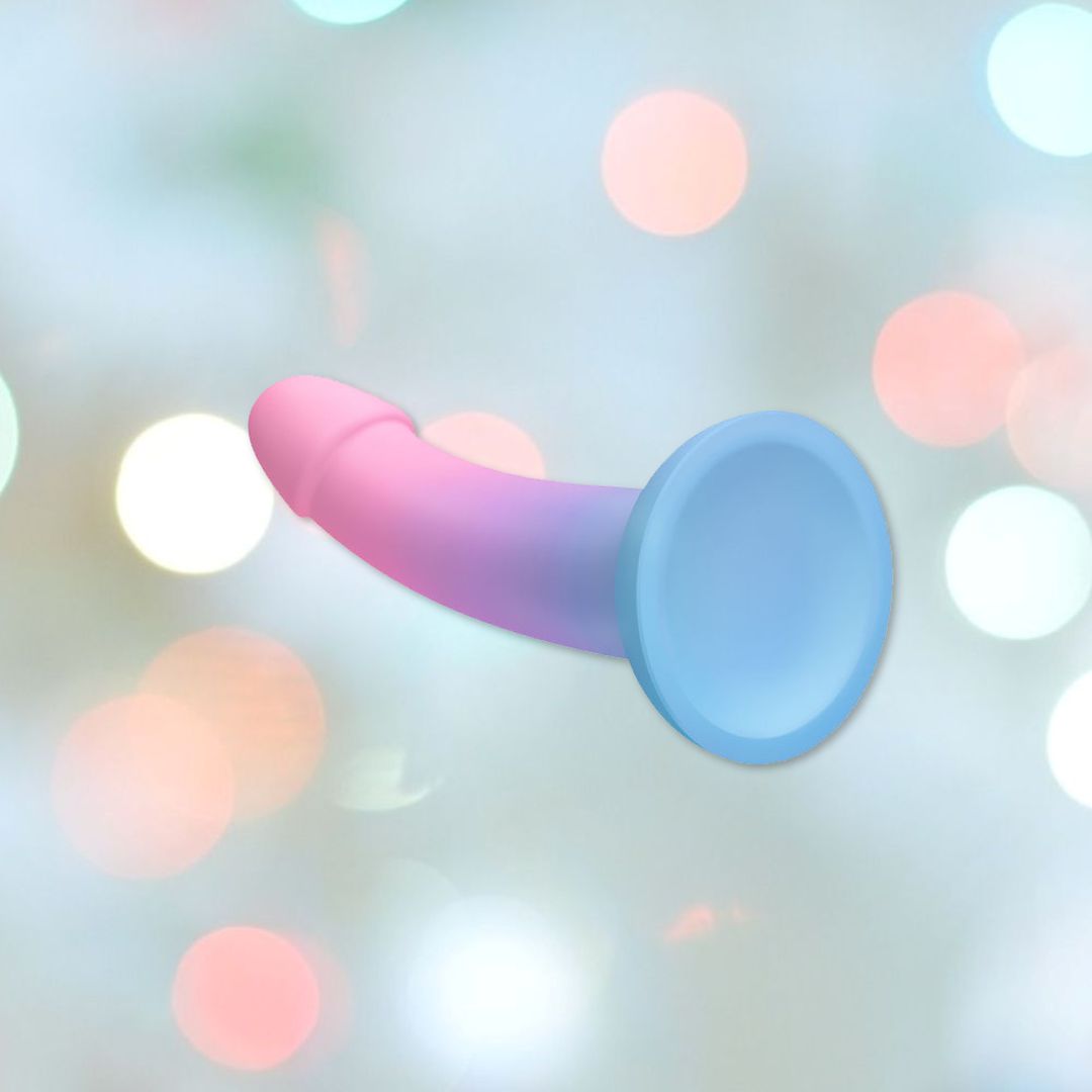 The Dildolls "Utopia" Silicone Dildo with Suction Base, featuring a sophisticated waterproof design and a stunning gradation of colors from pale pink to light blue, is shown against a blurred background adorned with soft white and pink bokeh lights.