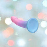 The Dildolls "Utopia" Silicone Dildo with Suction Base, featuring a sophisticated waterproof design and a stunning gradation of colors from pale pink to light blue, is shown against a blurred background adorned with soft white and pink bokeh lights.