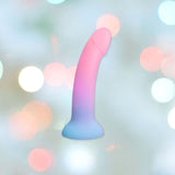 The Dildolls "Utopia" Silicone Dildo with Suction Base, a harness-compatible, phallic-shaped object with a gradient color scheme of pink to blue, is displayed against a soft, out-of-focus background featuring circular light bokeh in various pastel colors.