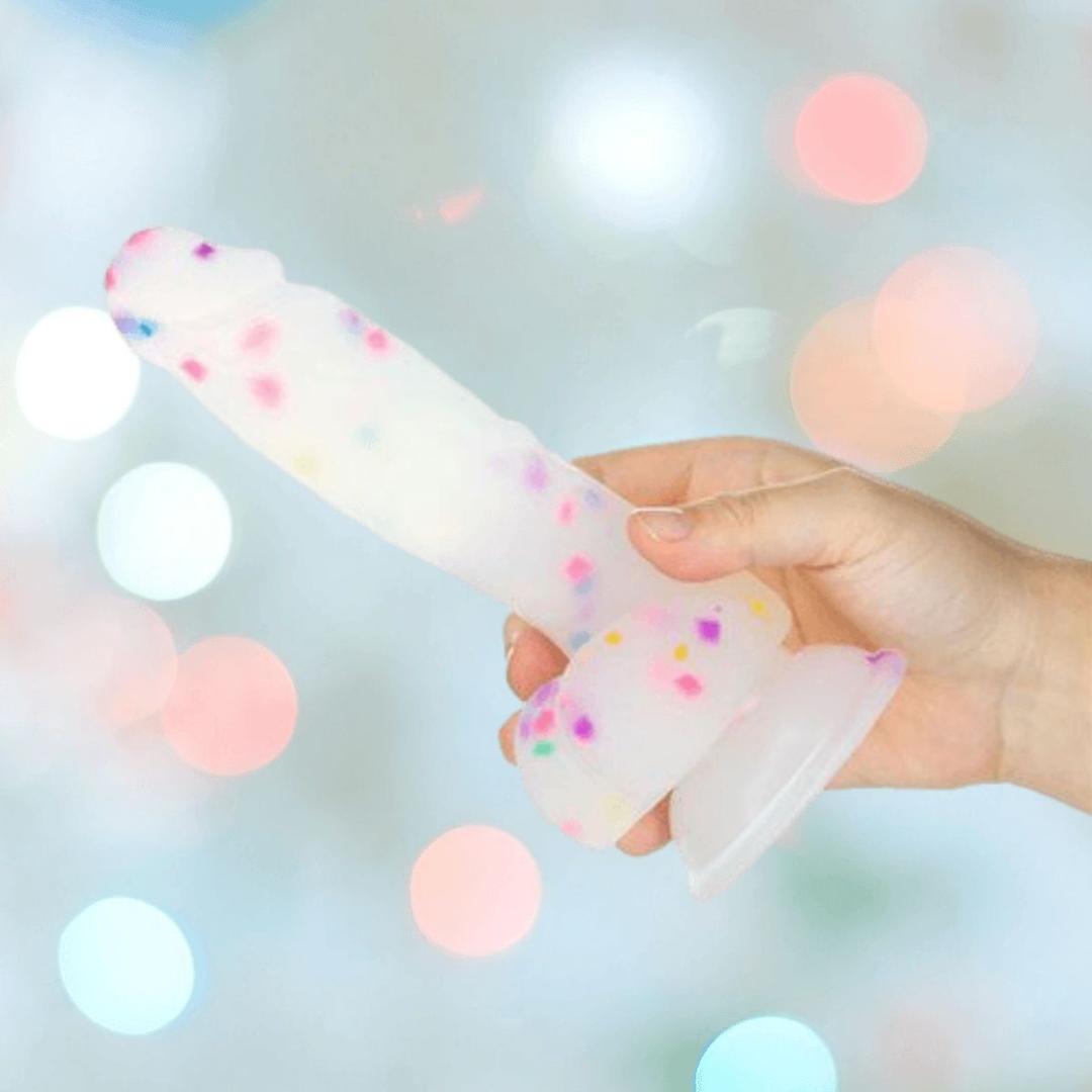 A hand holds the BMS Addiction Party Marty Vibrating Dildo - Rainbow 7.5", a white silicone toy with rainbow confetti and a suction base, against a blurred pastel background.