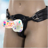 A person wears a black harness with the vibrant, speckled Addiction Party Marty Vibrating Dildo - Rainbow 7.5" by BMS; the softly blurred background enhances the colorful effect.
