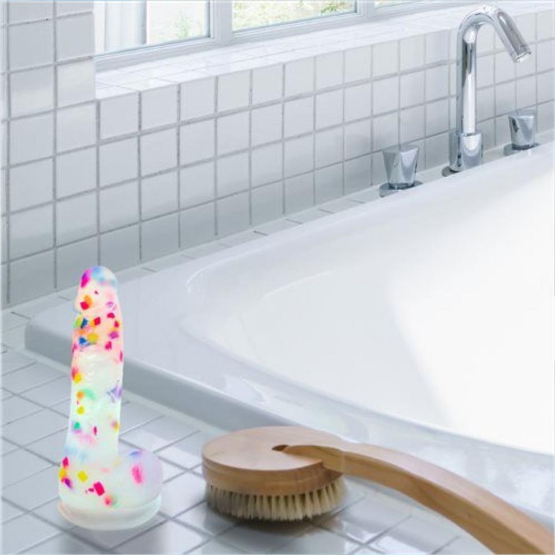 The BMS Addiction Party Marty Vibrating Dildo - Rainbow 7.5" rests on a white tiled bathtub's edge beside a wooden bath brush, with a silver faucet and window visible in the background.
