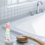 The BMS Addiction Party Marty Vibrating Dildo - Rainbow 7.5" rests on a white tiled bathtub's edge beside a wooden bath brush, with a silver faucet and window visible in the background.