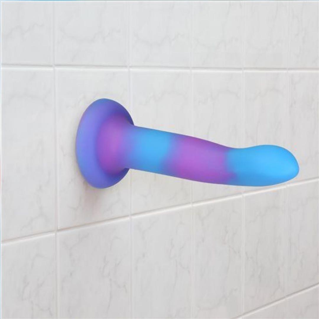 The Addiction Rave Glow-in-the-Dark Bendable Vibrating Dong by BMS in blue and purple (8") is attached to a white tiled bathroom wall.