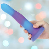 A hand holds the BMS Addiction Rave Glow-in-the-Dark Bendable Vibrating Dong - Blue Purple 8", featuring a strong suction base, against a softly blurred background with pastel-colored lights.