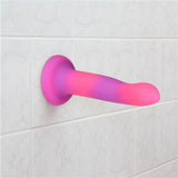 A pink and purple Addiction Rave Glow-in-the-Dark Bendable Vibrating Dong by BMS is attached to a white tiled bathroom wall using its suction base.