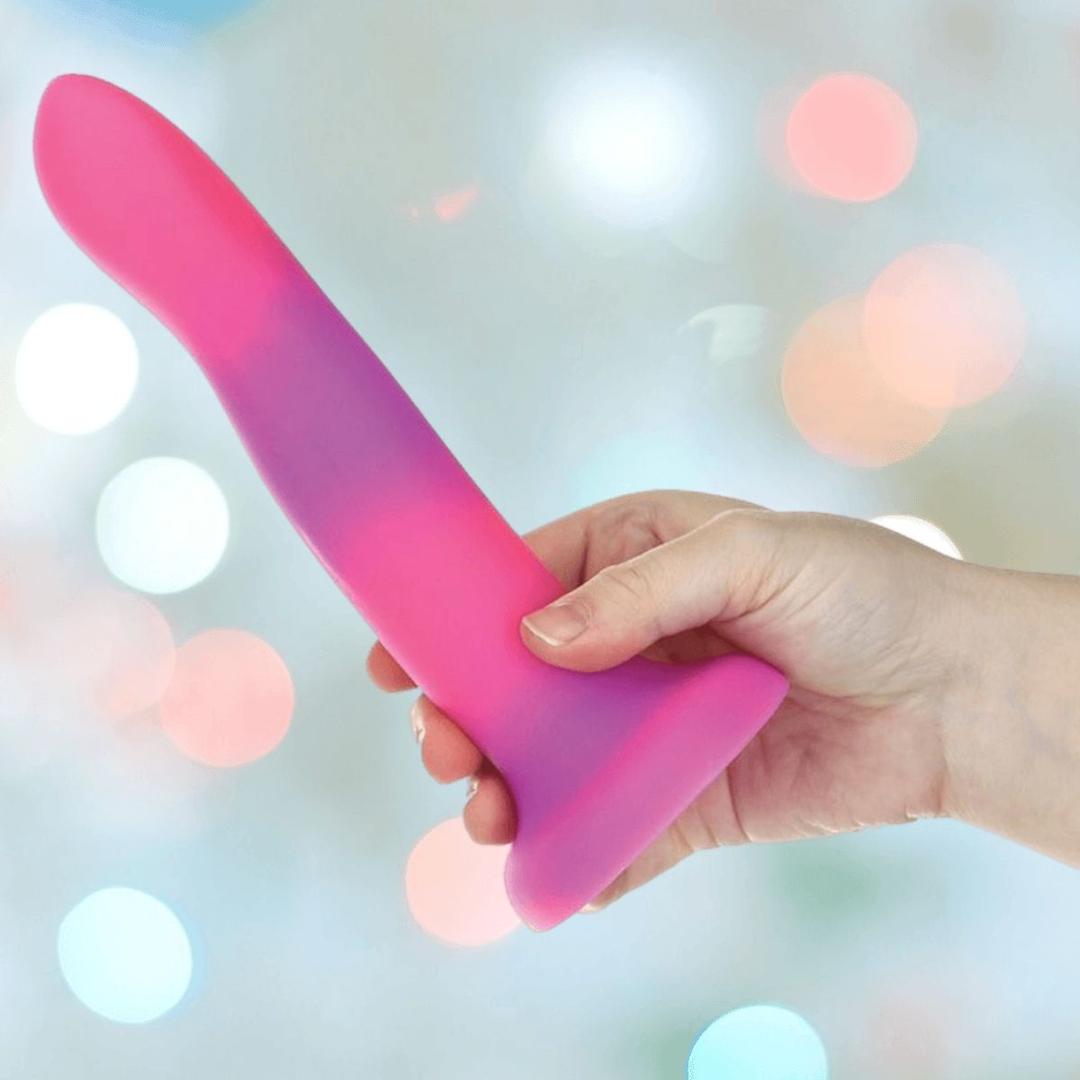 A hand holds the BMS Addiction Rave Glow-in-the-Dark Bendable Vibrating Dong, Pink Purple 8", featuring a strong suction base, against a softly blurred, colorful background.