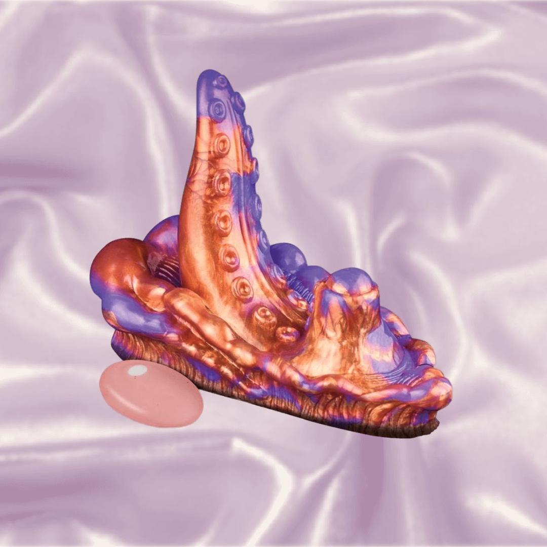 The Icon Brands Alien Nation Copperhead Grinder by Icon Brands, with its vibrant, iridescent finish and abstract tentacle-like shape, rests on silky lavender fabric. A smooth pink stone is positioned nearby for added visual intrigue.