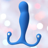 The Aneros Maximus Syn Trident Series Special Edition is a blue silicone prostate massager, featuring a bulbous top and two flexible arms with loops, displayed against a soft pastel pink and white bokeh background.