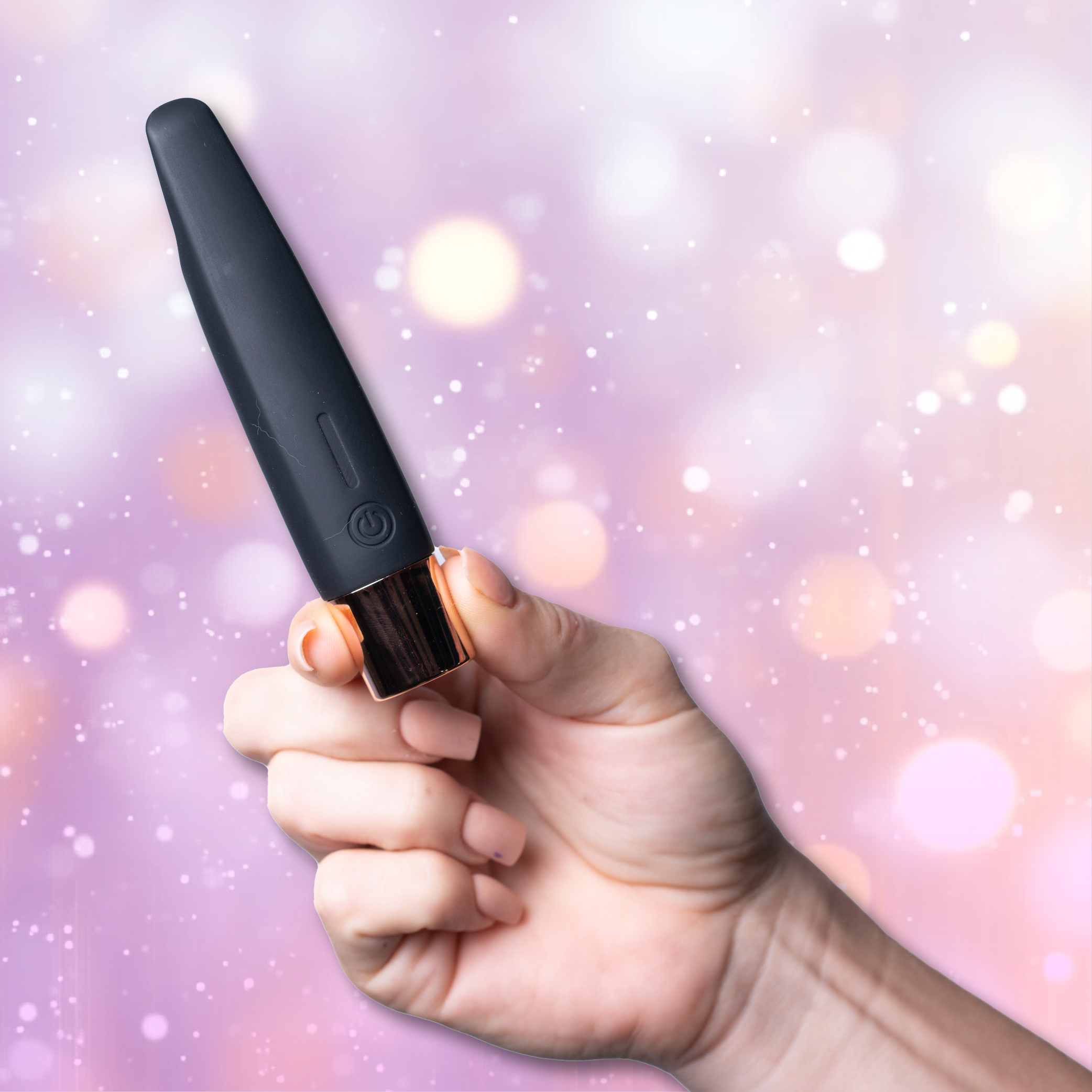 A hand holds the Aspen 15-Function Flickering Tip Vibrator by Maia Toys, featuring a sleek black design with a gold accent, set against a soft pastel bokeh background.