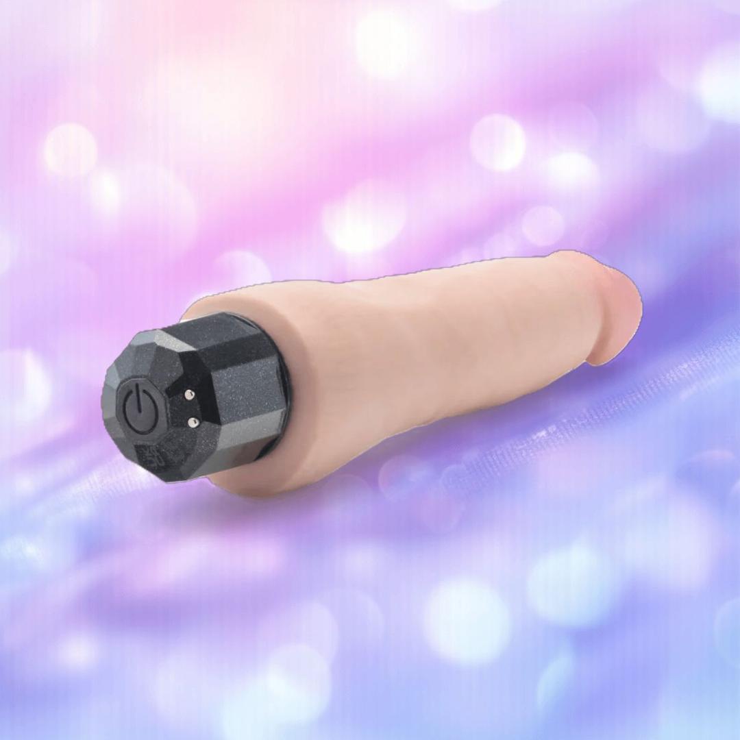The Au Naturel Vanilla Ice Vibrating Dildo by Blush Novelties, with a lifelike skin tone and black faceted control cap, rests on a soft, pastel purple and blue bokeh background.