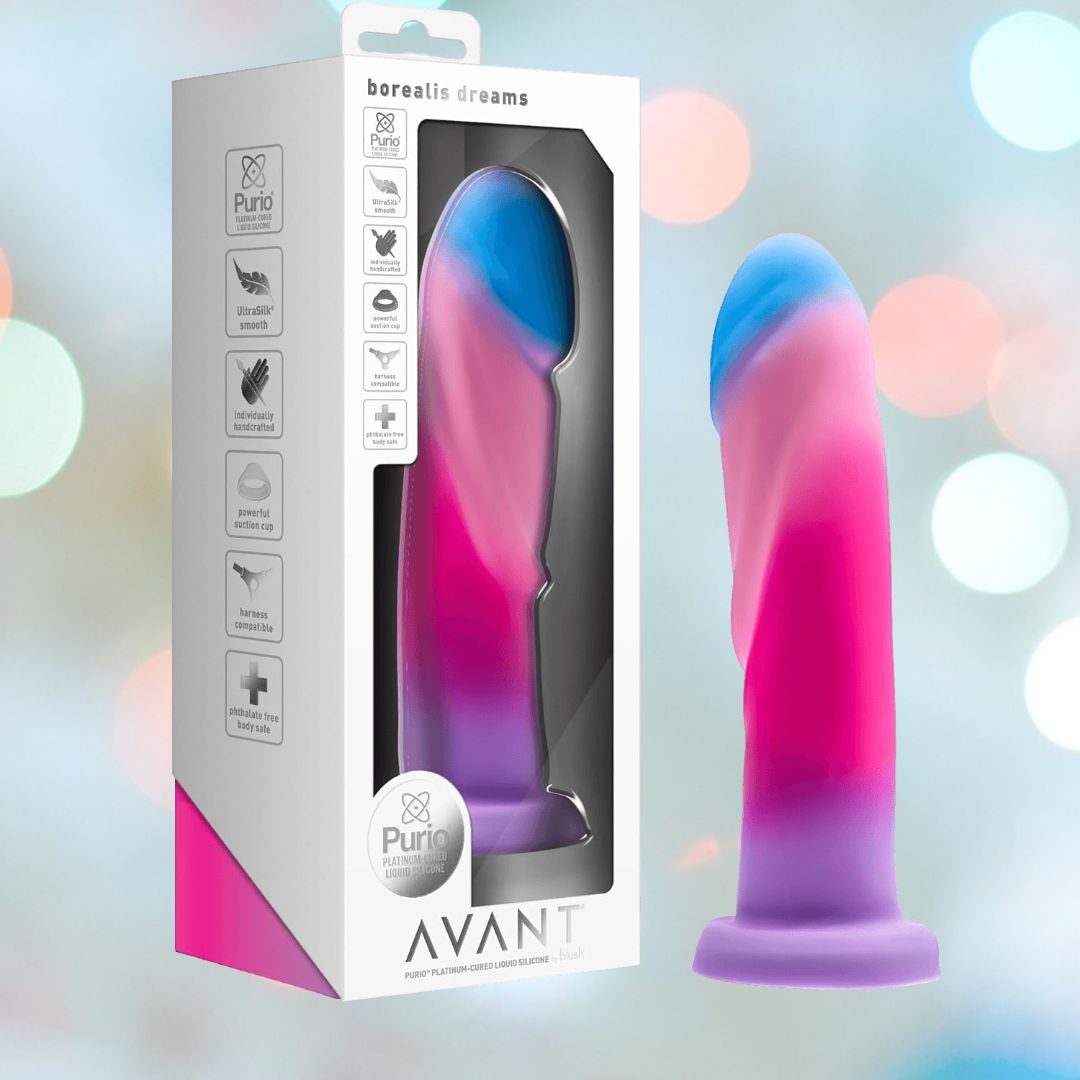 The packaging of the Avant Borealis Dreams - Cotton Candy Realistic Dildo displays a vibrant swirl of pink, purple, and blue. It highlights a hand-sculpted design crafted from platinum-cured silicone and features a strong suction base set against a blurred pastel backdrop. Additionally, it's compatible with harnesses for versatile use.