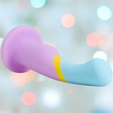 The Avant D14 G-Spot or Prostate Dildo - Heart of Gold by Avant is a pastel, platinum-cured silicone toy with pink, yellow, and blue sections, featuring a curved shape against a blurred background with colorful light bokeh.