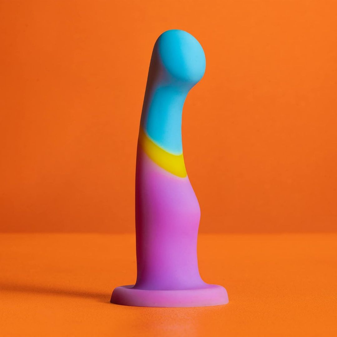 The Avant D14 G-Spot or Prostate Dildo – Heart of Gold by Avant, a colorful platinum cured silicone toy with a curved shape in blue, yellow, and purple, stands upright on an orange surface against an orange background.