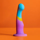 The Avant D14 G-Spot or Prostate Dildo – Heart of Gold by Avant, a colorful platinum cured silicone toy with a curved shape in blue, yellow, and purple, stands upright on an orange surface against an orange background.