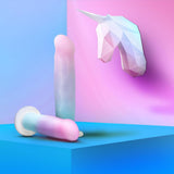 Two pastel Avant D17 'Lucky' Non-Porous Silicone Dildos by Avant rest on a blue surface, while a geometric unicorn sculpture pops against a pink and blue gradient wall, creating a whimsical, fantasy-inspired setting.