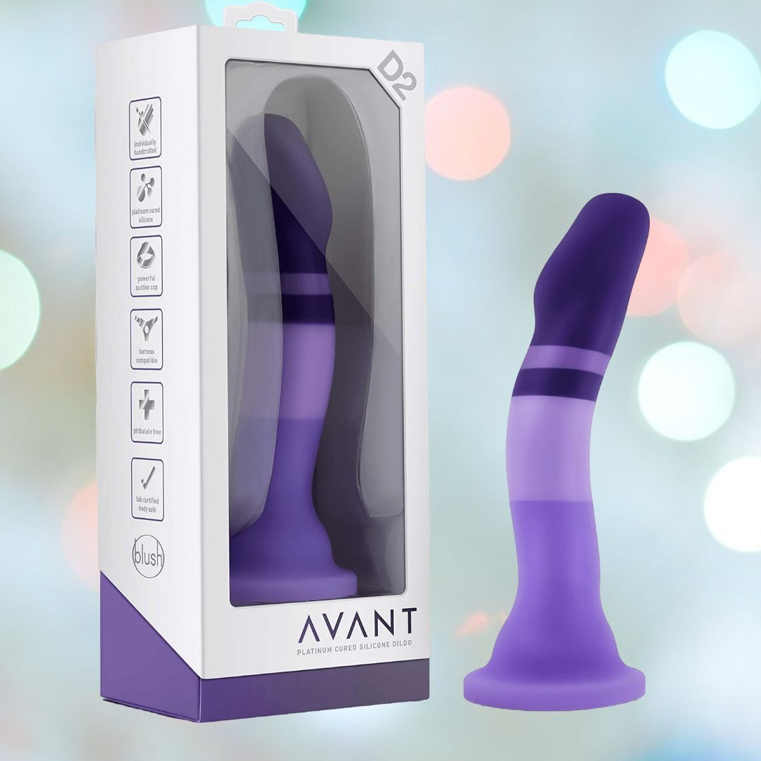 The boxed product titled "Avant D2 6" Non-Porous Silicone Dildo - Purple Rain" is displayed next to the identical unboxed item. This silicone dildo has a curved, cylindrical shape in various shades of purple and includes a suction cup base. The packaging showcases icons that indicate its features and provides brand information for Avant.