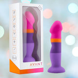 The Avant D3 7" Non-Porous Silicone Dildo - Summer Fling, featuring vibrant shades of pink, orange, and purple with a suction cup base, is showcased alongside its packaging. The box highlights the product's features and displays the brand name "Avant." A soft background with blurred lights emphasizes the body-safe design.