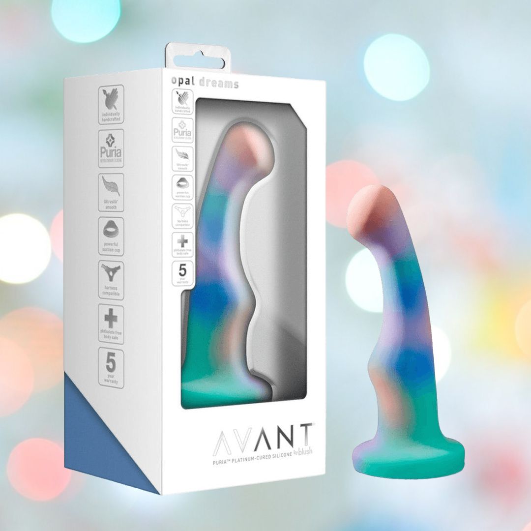 An aqua Avant Opal Dreams Dildo stands next to its white box, which displays product details and a clear window showing the toy. The softly blurred background glows with pastel lights.