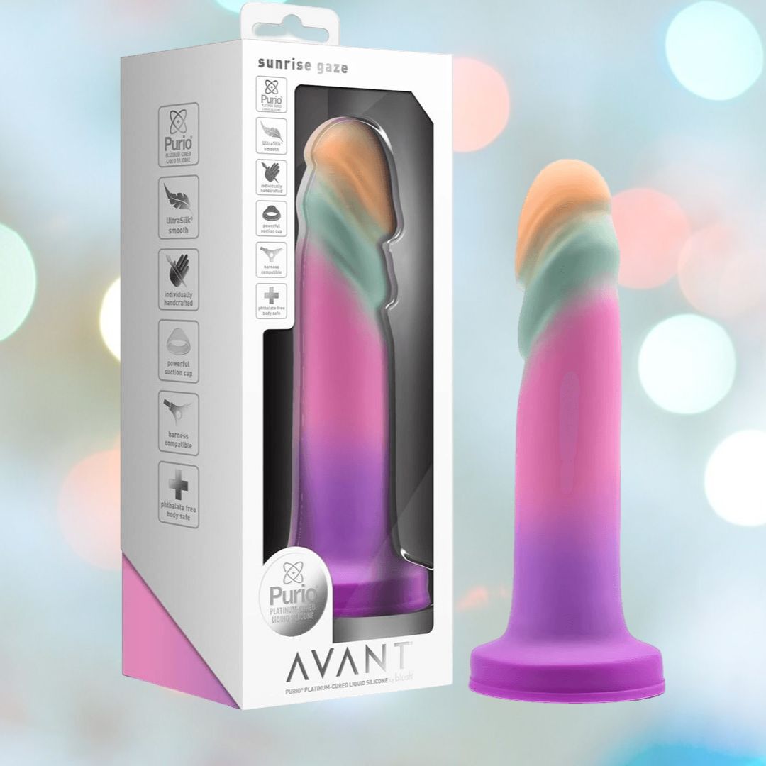 A vibrant artisanal dildo in shades of pink, purple, and light blue is showcased in packaging labeled "Avant Sunrise Gaze - Sherbet Realistic Dildo" from the Avant brand. It features a platinum-cured silicone construction and a suction cup base, set against a backdrop of soft pastel bokeh lights for a dreamy effect.