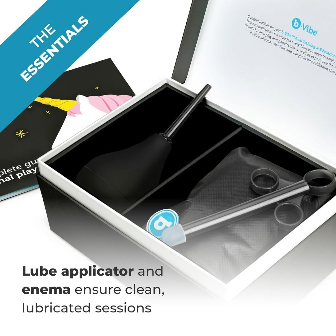 The open box labeled "B-Vibe Anal Training Set of 3 Butt Plugs - Black" includes a lube applicator and enema, as well as an anal training kit. Inside, a black bulb with a nozzle and a clear applicator tube are neatly arranged. A card featuring a unicorn illustration is partially visible.