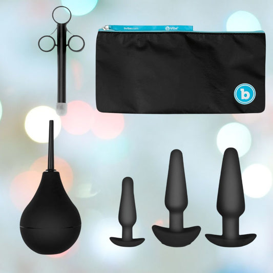 A set of B-Vibe black silicone adult products, the Anal Training Set includes a small, medium, and large silicone butt plug along with a bulb and cleaning tool. Each item is displayed on a blurred, colorful background with a black zippered pouch positioned above them. 1080