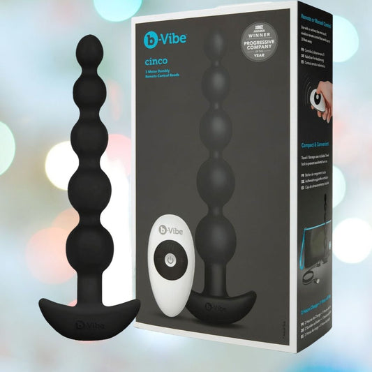 The B-Vibe Cinco Beads XL Vibrating Anal Beads in black, its remote control, and the branded packaging are shown against a softly blurred pastel background. 1080