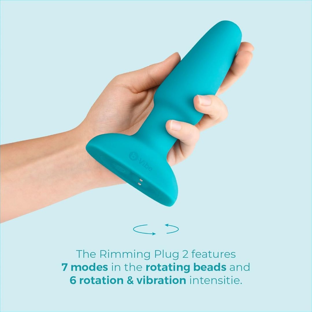 A hand holds the B-Vibe Rimming Plug 2 Vibrating Butt Plug by B-Vibe. Text below lists its features: 7 rotating bead modes, 6 vibration and rotation intensities, and a wireless remote for easy control.