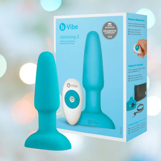 A blue B-Vibe Rimming Plug 2 Vibrating Butt Plug is shown with a white remote beside its packaging, featuring the product image and highlights from B-Vibe against a soft blue background. 1080