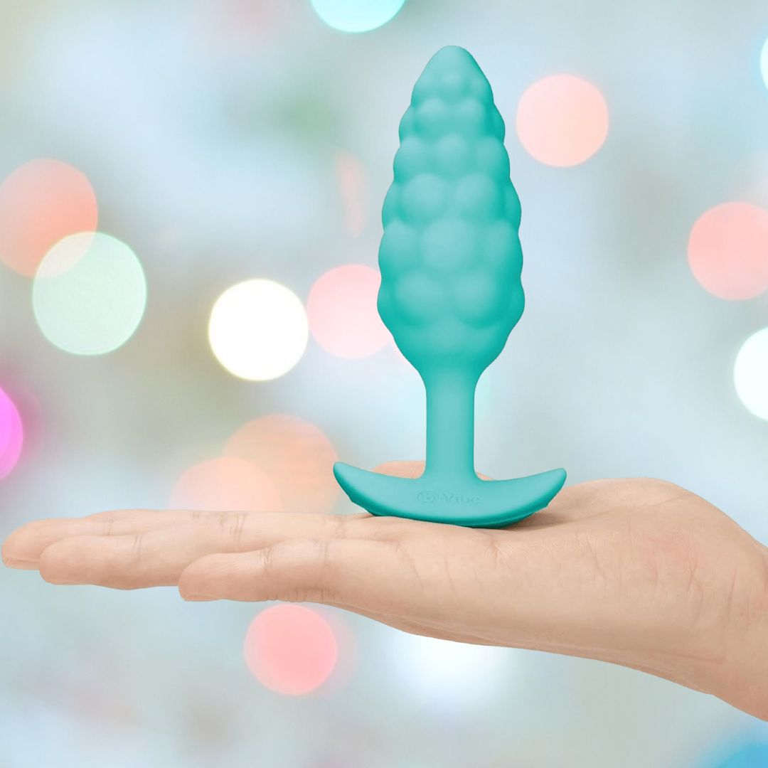 A hand holds the B-Vibe Texture Vibrating Butt Plug Bump - Mint (Small), a teal, body-safe silicone toy with a flared base by B-Vibe, set against a soft pastel background with circular light bokeh.
